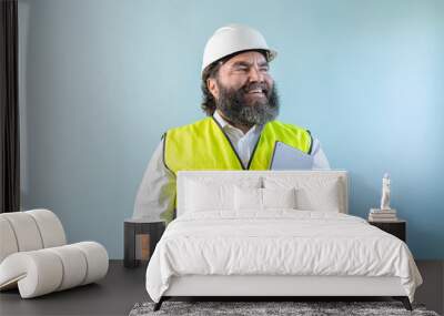 smiling adult man engineer with beard and wearing helmet and safety vest looking to the right holding his touch pad on blue background Wall mural