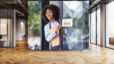 Happy waitress standing at restaurant entrance. Portrait of african american business woman attend new customers in her coffee shop. Happy woman owner showing open sign in her small business shop. Wall mural