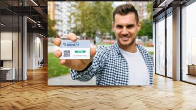 Happy young man showing health passport indicating that he is immune to coronavirus. Selective focus on card in foreground Wall mural