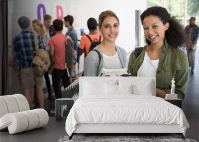 Portrait of two smiling female students holding books, other students  Wall mural