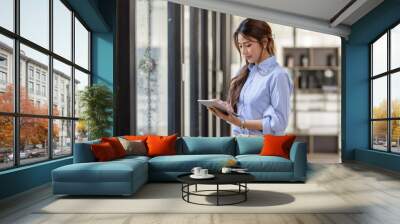 Young business Asian woman using tablet ipad, standing near the window in workplace Wall mural