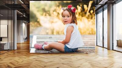 Little blond-haired girl sitting on a concrete platform in the field at sunset Wall mural