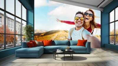 Cheerful couple of hikers taking selfie on top of the mountain - Millennial guy and girl enjoying summertime day out laughing at camera together - Millenial travelers standing on nature background Wall mural