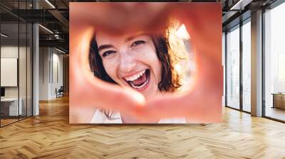 Close up image of smiling woman in swimwear on the beach making a heart shape with hands - Pretty joyful woman laughing at camera outside - Healthy lifestyle, self love and body care concept Wall mural