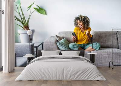 Excited happy young black woman holding smart phone device sitting on sofa at home - Happy satisfied female looking at mobile smartphone screen gesturing yes with clenched fist - Technology concept Wall mural