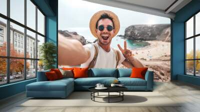 Handsome man wearing hat and sunglasses taking selfie picture on summer vacation day - Happy hiker with backpack smiling at camera outside - Tourist walking on the beach - Traveling and technology Wall mural