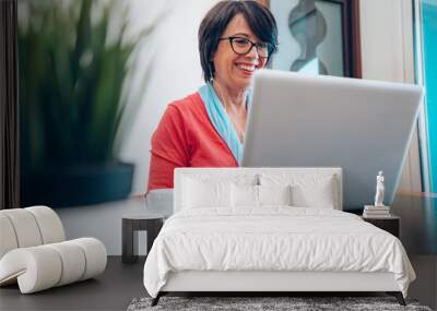 Mature woman using pc laptop indoor - Senior female smart working with computer at home - She is sitting on a wooden modern desk at home. Smiling and enjoying communication Wall mural