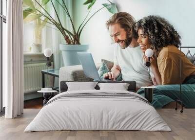 Multiracial young couple watching computer laptop sitting on the sofa at home - Happy diverse husband and wife using pc online services - Technology life style concept Wall mural