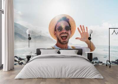 Young man with backpack taking selfie portrait outside - Smiling happy guy enjoying summer holidays at the beach - Millennial showing victory hands symbol to the camera - Youth and journey Wall mural