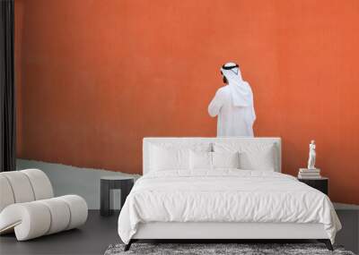 Arabic man with traditional emirates clothes walking outdoors in the street against a orange wall background - Arab ethnicity and muslim religion concept - Copy space for text Wall mural