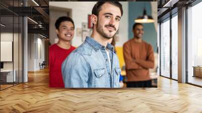 Portrait of young student looking at camera during leisure activity in recreation room at campus - Multi-ethnic classmates in background - Square cut with focus on the face Wall mural