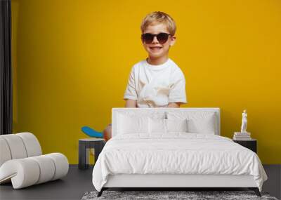 Adorable little kid wearing casual outfit and sunglasses smiling while sitting on skateboard with cross legged against yellow background. Wall mural