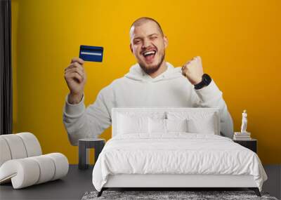 Excited lucky handsome man wear white hoodie rising fist while holding bank card isolated over yellow background Wall mural