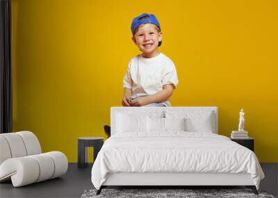 Full body length excited kid boy wearing trendy blue cap and white t-shirt, smiling while sitting on modern skateboard against yellow background. Wall mural