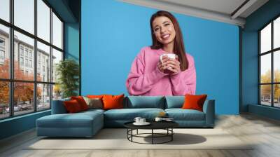 Happy young Hispanic woman in pink jumper smiling and holding cup of hot beverage while standing against blue background. People, emotions concept Wall mural