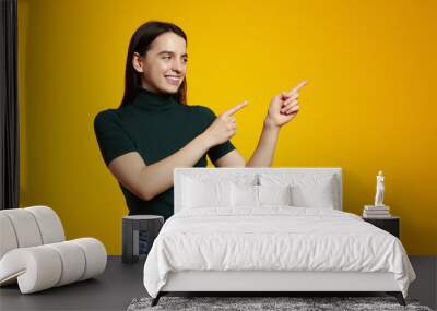 Photo of impressed brunette lady looking and pointing to promo wearing green t-shirt isolated on yellow color background Wall mural