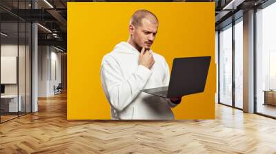 Portrait of a doubtful bearded man in white hoodie holding laptop computer while touching chin with hand isolated over yellow background Wall mural