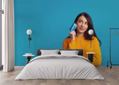 Thoughtful young girl wearing yellow clothes holding plastic credit card near face and looking at free empty space, isolated over blue background  Wall mural