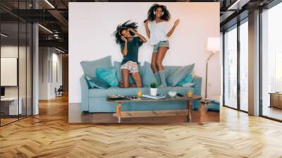 Happy young two black women sitting and standing up in the couch on the computer and mobile phone with headphones dancing . Wall mural