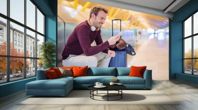 Young man waiting listening music and using mobile phone at the airport with a suitcase. Wall mural