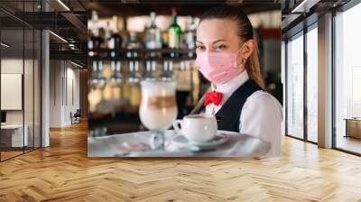 A female Waiter of European appearance in a medical mask serves Latte coffee. Wall mural