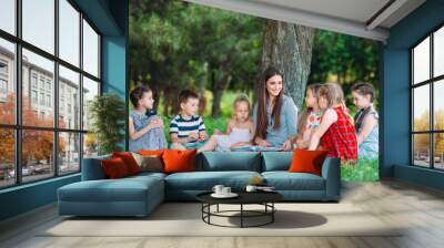 Children and education, young woman at work as educator reading book to boys and girls in park. Wall mural