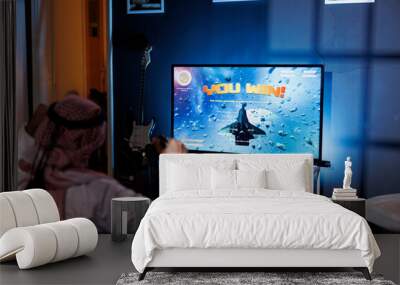 A young Arab man plays an immersive virtual reality game at home, celebrating victory while holding a wireless controller. He is sat at home, absorbed in amusement, and lighted by the screen light. Wall mural