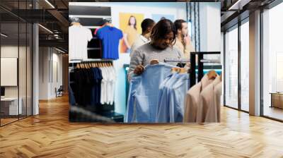 African american woman searching apparel size stock in clothing store online app using smartphone. Shopping mall customer holding mobile phone and checking garment on website Wall mural