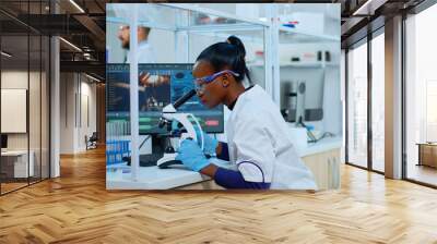 African scientist checking sample of virus using microscope in modern lab. Multiethnic team examining vaccine evolution using high tech for scientific research of treatment development against covid19 Wall mural