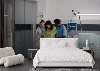 Afro american nurse with doctor helping patient to stand up from bed holding intravenous IV fluid drip bag after medical surgery in hospital ward. Practitioner monitoring sickness recovery Wall mural