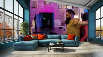 Bearded man sitting on gaming chair stretching while playing video games Wall mural