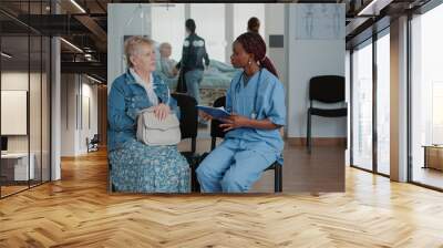 Close up of african american nurse talking to elder patient in waiting area. Medical assistant explaining disease and healthcare treatment to retired woman in waiting room at facility. Wall mural