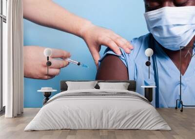 Close up of black nurse getting vaccinated with syringe and needle, wearing face mask. African american medical assistant receiving vaccine shot from hands of doctor for virus protection Wall mural