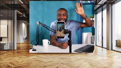 Closeup of live vlog setup with smarthone on stand filming content creator waving hand to followers in online morning show. Selective focus on vlogger saying hello at audience sitting at desk. Wall mural