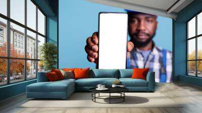 Confident african american contractor holding cell phone with white screen mockup design wearing coveralls in studio. Builder using touchscreen device for advertising construction industry concept. Wall mural