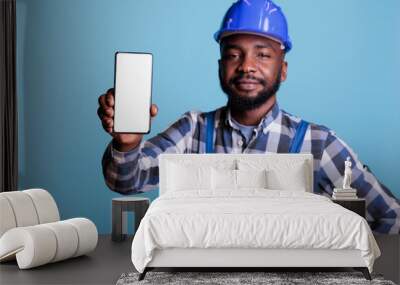 Construction worker in blue uniform work coveralls holds cell phone with empty screen for advertising isolated on blue background. Male professional builder in hard hat looking at camera. Wall mural