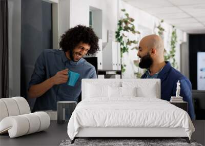 Corporate colleagues telling joke and laughing while drinking coffee in coworking space. Smiling young arab start up company employees enjoying work break in business office Wall mural