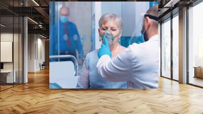 Doctor in mask and visor helping old lady to breathe with a respiratory oxygen mask while sitting on hospital bed in modern private clinic. Coronavirus covid-19 outbreak healthcare crisis. Wall mural