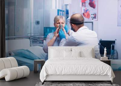 Doctor putting an oxygen mask on old retired senior woman during COVID-19 coronavirus outbreak in modern private hospital or clinic. Fighting infection and disease medicine and quarantine Wall mural