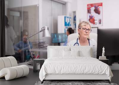 Elderly aged woman doctor working on pc in hospital office, Young therapist discussing with invalid patient in wheelchair and nurse looking at x-ray on computer screen. Wall mural