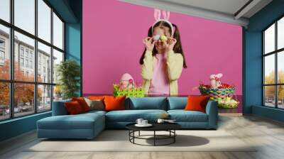 Enthusiastic young girl playing peek a boo in front of camera, using painted easter eggs against pink background. Joyful lovely toddler feeling excited about spring holiday festivity. Camera B. Wall mural