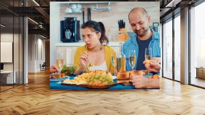 Extended family interacting while having a meal at dining table in the kitchen. Multi generation, four people, two happy couples talking and eating during a gourmet dinner, enjoying time at home. Wall mural