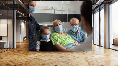 Family wearing protective face mask against coronavirus while visiting elderly senior woman during clinical recovery in hospital ward. Doctor woman discussing healthcare treatment. Medkcal diagnosis Wall mural