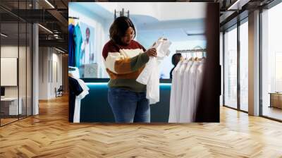 Fashion boutique client holding white shirt on hanger and checking fabric quality. African american woman examining apparel rack while searching for formal outfit in clothing store Wall mural