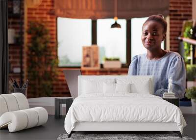 Female remote worker sitting near open laptop and looking at camera in modern home office. Young successful african american businesswoman at workplace in contemporary room Wall mural