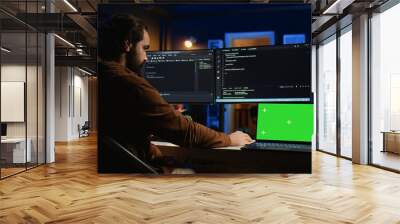 Green screen laptop next to freelancing man doing IT support job from apartment office, coding on PC. Mockup notebook on desk where software engineer writes lines of code on computer at home, camera B Wall mural