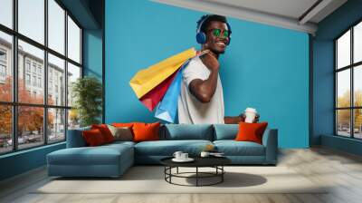 Happy man with handbags dancing after shopping spree. Cheerful person with sunglasses listening to music on headphones while holding bags with clothing for sale purchase on black friday Wall mural