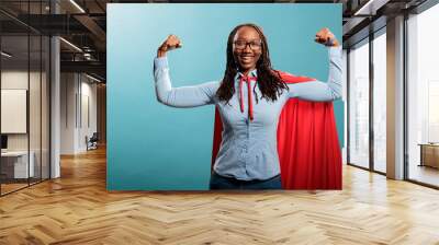 Happy positive justice defender flexing arms as a sign of strength while standing on blue background. Proud and tough looking young adult superhero woman showing empowerement and braveness. Wall mural