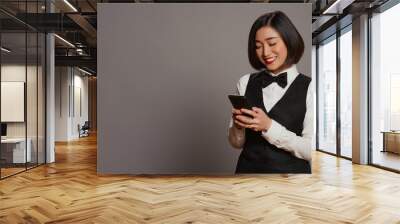 Hospitality industry worker using smartphone app over grey background, looking at online information to confirm room reservations. Asian receptionist browses websites. Camera B. Wall mural