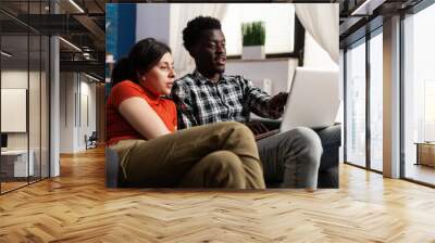 Interracial couple looking at modern laptop computer in living room. African american man holding digital gadget while caucasian woman watching device screen. Mixed race people Wall mural